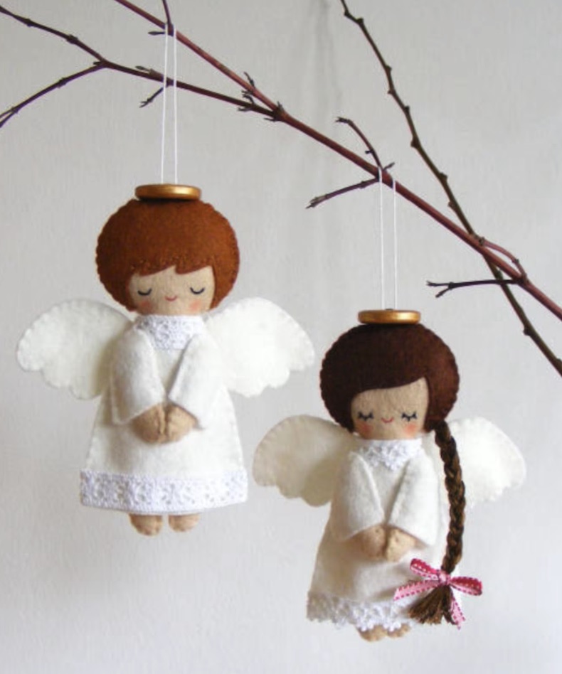 Two handmade felt angel ornaments with brown hair and white dresses hang from a branch. One has short hair; the other has a long braid tied with a ribbon. Both have small gold halos.