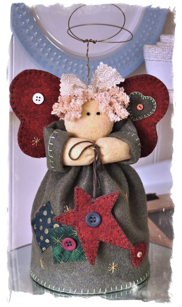 A handmade fabric angel doll with red felt wings, curly hair, and button decorations on its green dress, holding a cord, with a wire halo above its head.