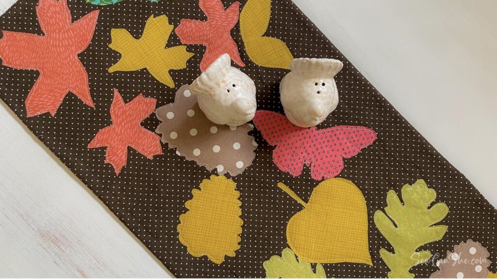 Two ceramic bird-shaped salt and pepper shakers sit on a brown table runner decorated with colorful fabric leaves and a white polka dot pattern.