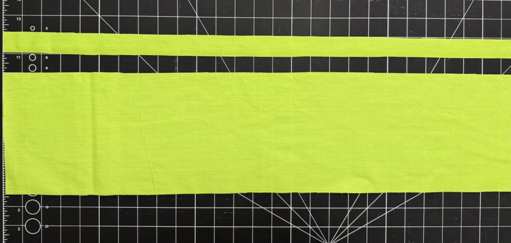 Two strips of bright yellow-green fabric, perfect for crafting an advent tree, one narrow and one wide, are laid horizontally on a black grid cutting mat.