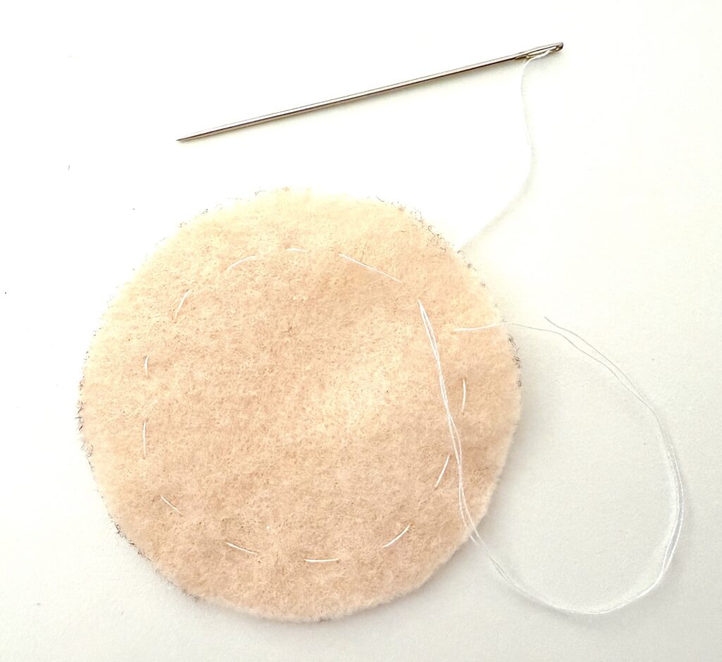A round piece of light-colored felt with a needle and white thread partially stitched around its edge lies on a white background.
