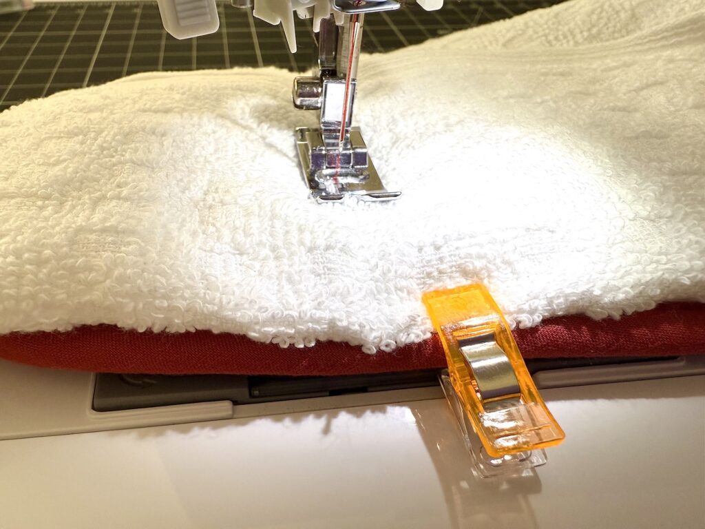 A sewing machine stitches layers of white and red fabric together, with an orange fabric clip holding the materials in place.