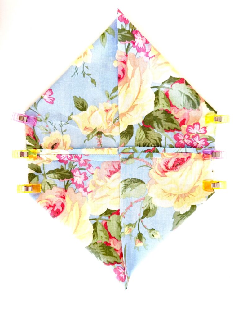 A floral fabric square with folded corners, resembling a tissue box cover, is held in place by four colored clips on a white background.