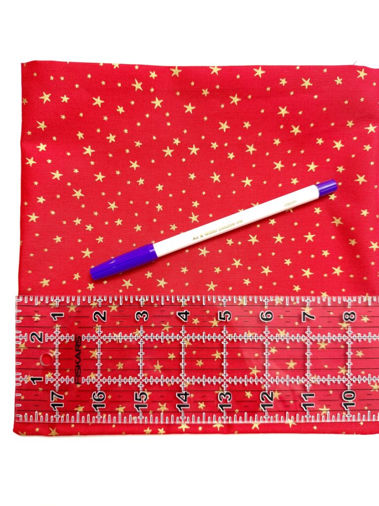 A clear ruler and a blue pen rest on a red fabric with a gold star pattern. The ruler shows measurements in inches, and the fabric is laid flat.