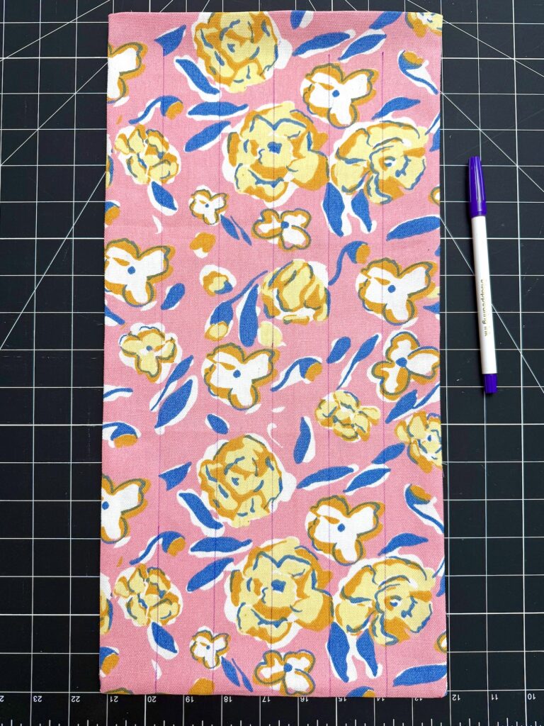 A rectangular piece of pink fabric with yellow and white floral patterns, possibly intended as a trivet, is placed on a cutting mat next to a purple and white pen for scale.