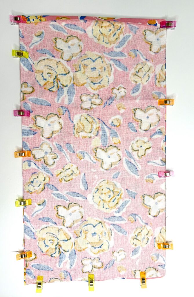 A rectangular piece of pink floral fabric, perfect for a trivet project, is secured along the edges with multicolored sewing clips on a white background.