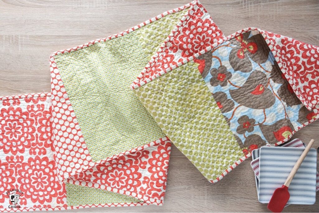 Four quilted placemats with various red, green, and brown floral and geometric patterns are displayed on a wooden surface with a striped napkin and a red spatula.