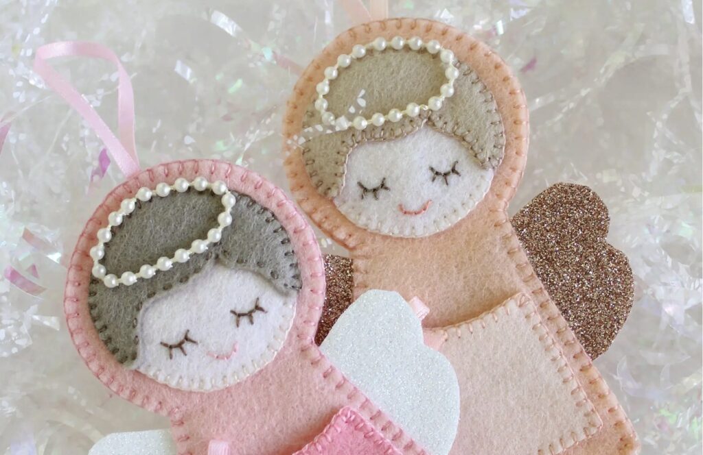 Two handcrafted felt angel ornaments with stitched faces, pearl bead halos, glittery wings, and pastel colors, lying on a shimmery background.