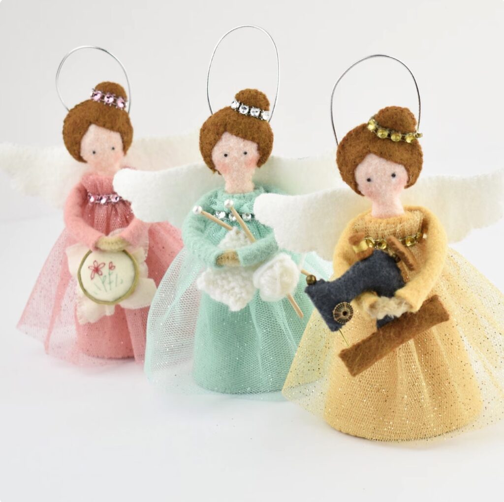 Three fabric angel ornaments with brown hair, each wearing a pastel-colored dress and holding sewing-themed items, are displayed against a white background.