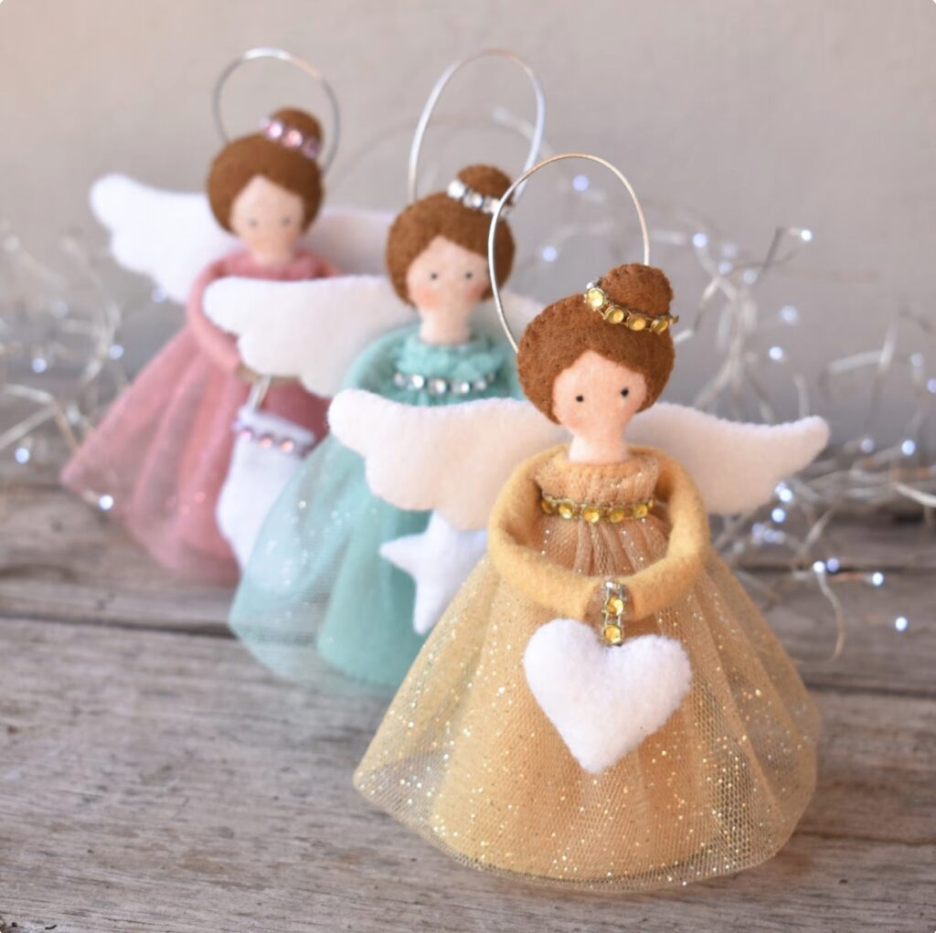 Three handmade felt angel ornaments with wings and sparkling dresses stand on a wooden surface, each holding a white heart. String lights are blurred in the background.