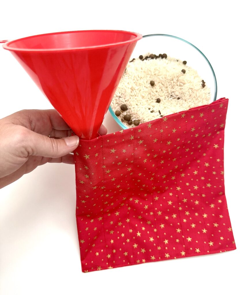 A hand holds a red funnel over a red fabric pouch with gold stars, next to a bowl of rice mixed with beans.