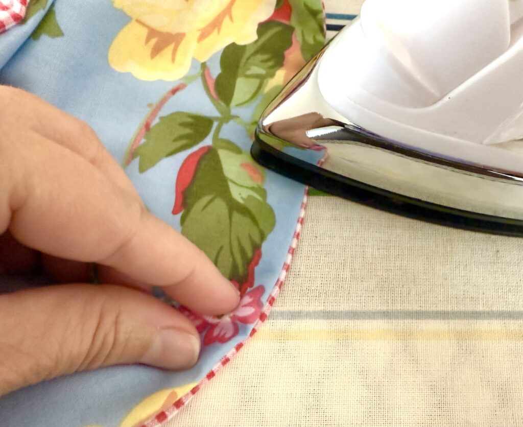 A hand holds the edge of a floral fabric, possibly crafting a tissue box cover, while an iron rests nearby on an ironing board.