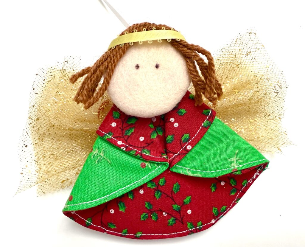 Handmade cloth angel ornament with a round fabric face, yarn hair, a gold headband, gold mesh wings, and a red and green dress with a holly pattern.