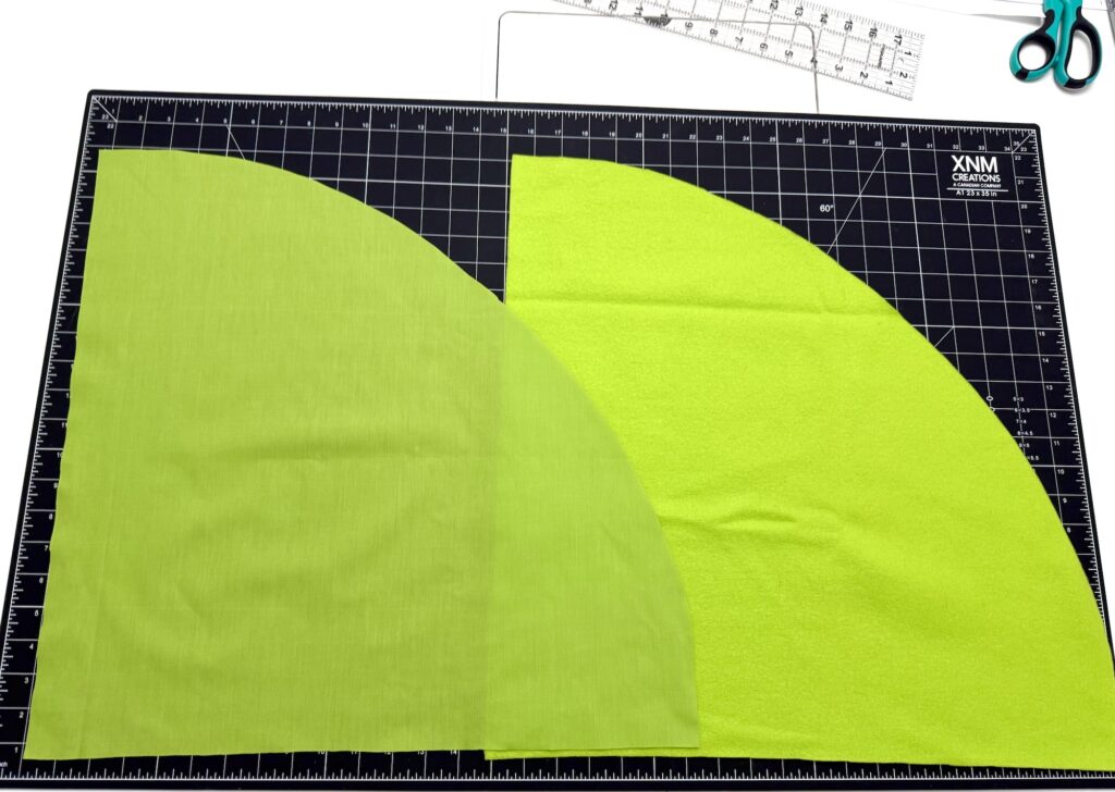 Two large quarter-circle pieces of green fabric, perfect for crafting an advent tree, are placed on a black grid cutting mat, with a ruler and scissors visible in the background.