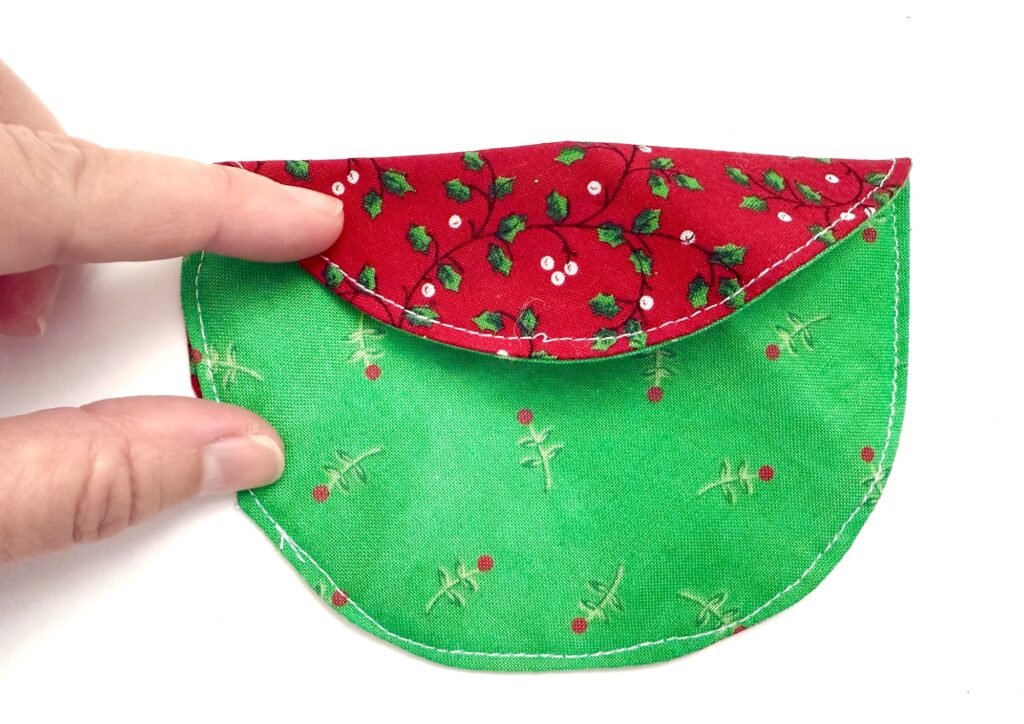 A hand holds a small, sewn fabric pouch with a red holly-patterned flap and a green body decorated with small red and white details.