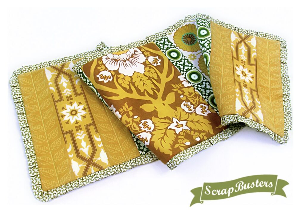 A fabric book cover made from yellow, green, and brown patterned scrap fabrics lays open, showing multiple vintage-style designs. A "ScrapBusters" label appears in the corner.