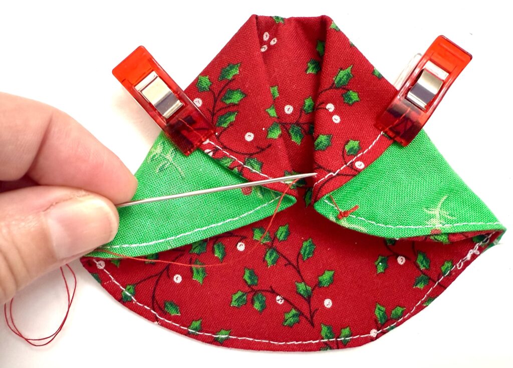 A hand sewing two layers of red and green fabric with a needle, with two red clips holding the fabric in place.