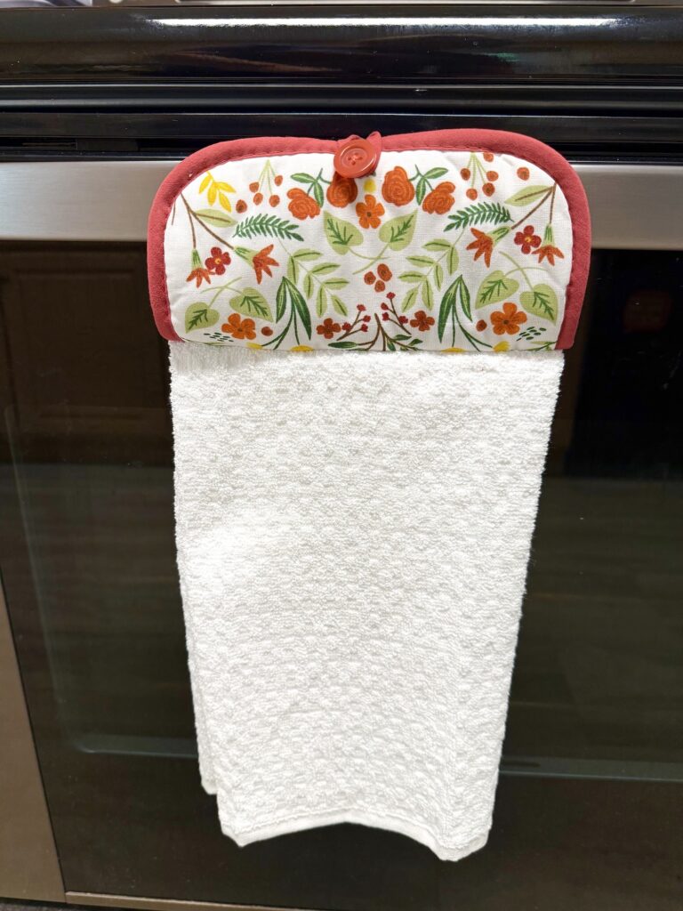 A white kitchen towel with a patterned fabric top featuring red and orange flowers is hanging from an oven handle, adding a cheerful touch to any kitchen décor.