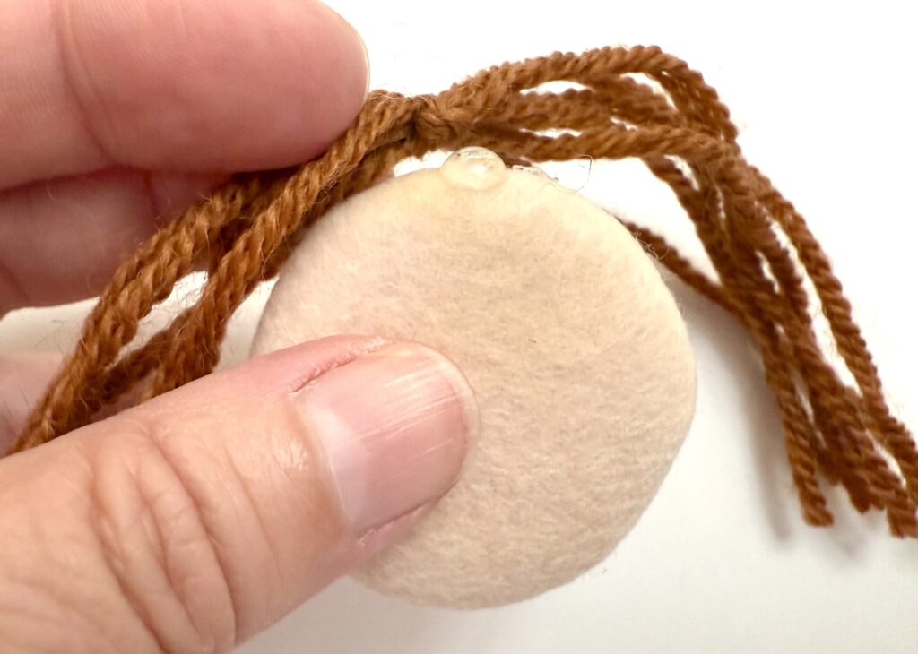 A hand holds a round beige felt piece with brown yarn strands glued to the top.