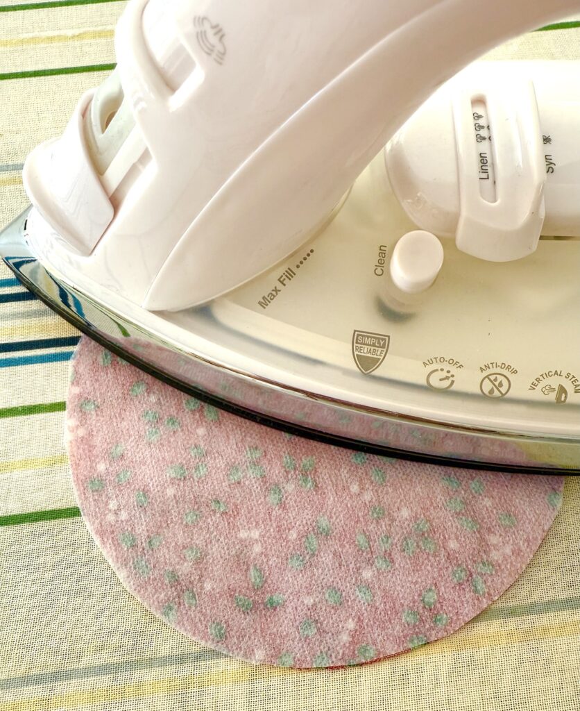 A white iron presses a pink fabric circle with small green dots on a striped ironing board cover.