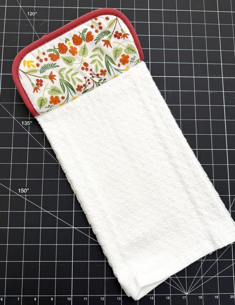 A handmade fabric towel with a floral-patterned top section and a plain white terry cloth bottom, placed on a black grid cutting mat.