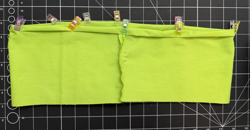 Two pieces of bright green fabric, perfect for crafting an advent tree, with folded edges pinned by multicolored clips, are placed on a black cutting mat with white measurement lines.