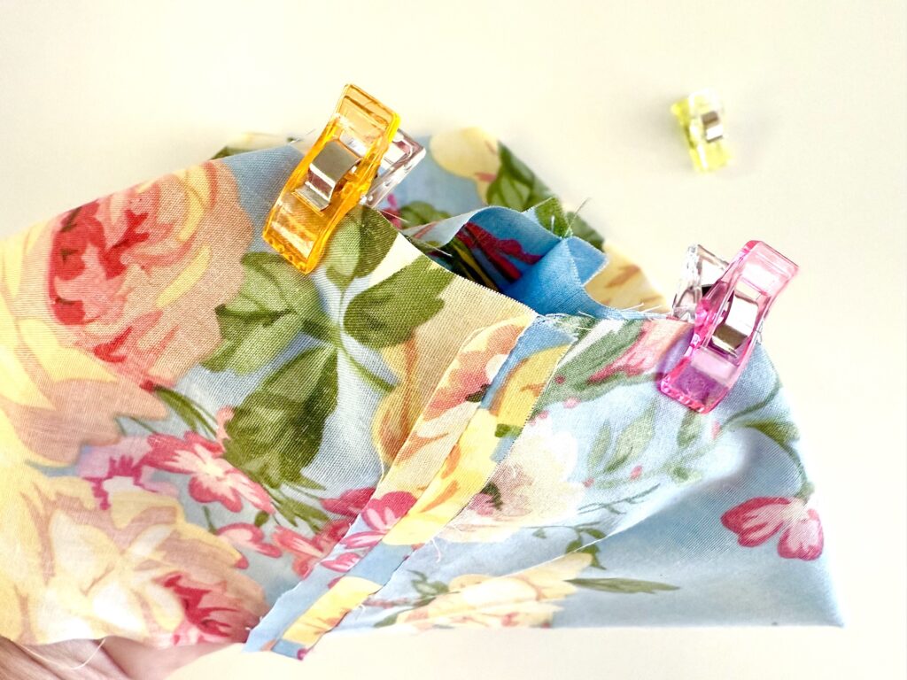 A piece of floral fabric, intended for a tissue box cover, is held together with colorful sewing clips on a white surface, with one clip positioned separately in the background.