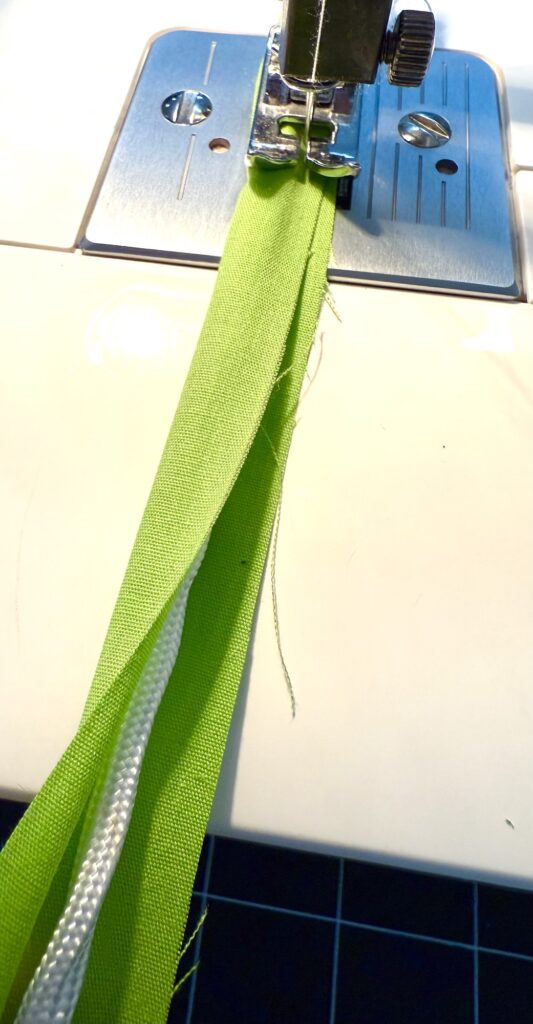 Close-up of a sewing machine stitching green fabric for an advent tree, with a white cord inserted, showing the needle, presser foot, and part of the sewing plate.