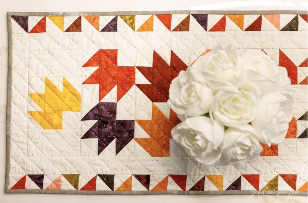 A white vase with white flowers sits on a quilted table runner featuring a geometric maple leaf pattern in autumn colors.
