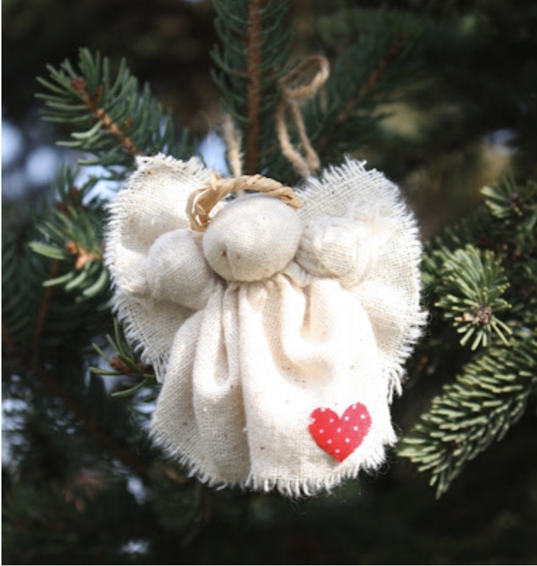 A handmade fabric angel ornament with a red heart detail hangs on a pine tree branch.