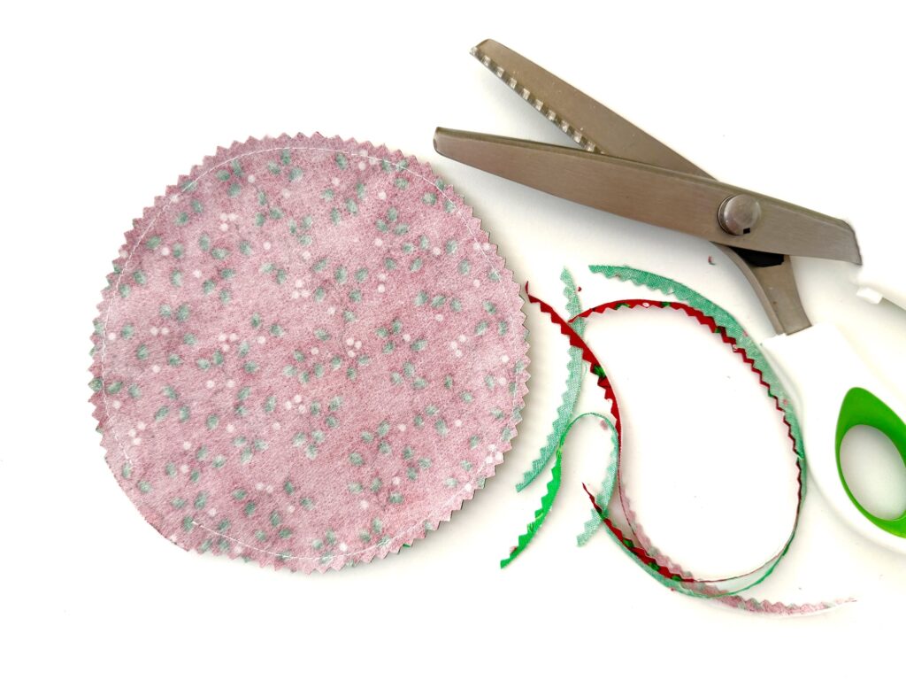 A round piece of pink fabric with a textured pattern, cut with pinking shears. The shears and scraps of red and green fabric are beside it on a white background.