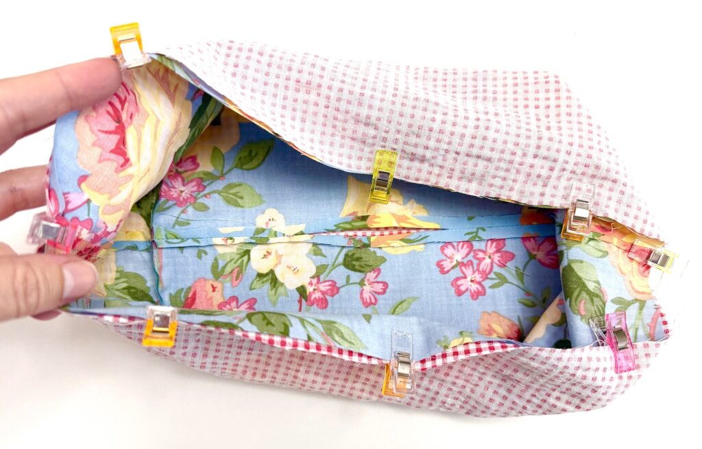 A hand holds a partially sewn fabric pouch with floral and checkered patterns, secured with colorful clips along the top edge—a charming tissue box cover in the making.
