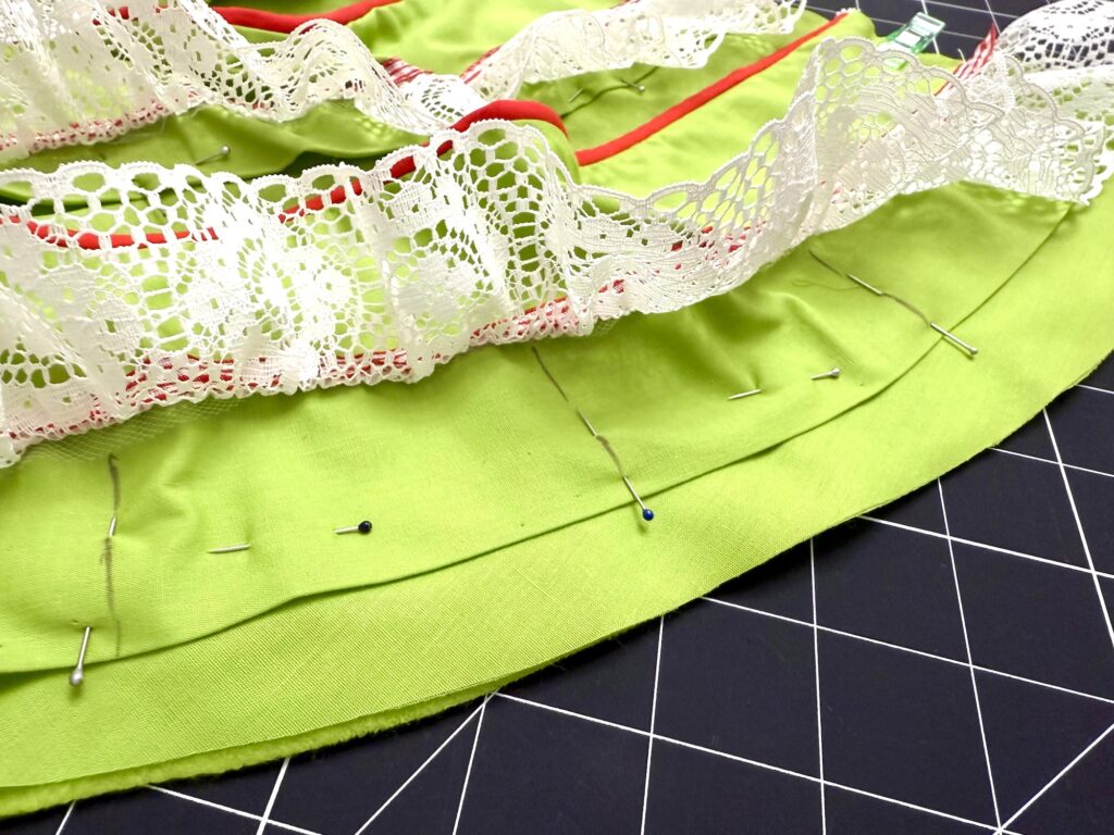 Green fabric with lace trim and red ribbon, perfect for crafting an advent tree, is pinned together with sewing pins on a black grid cutting mat.