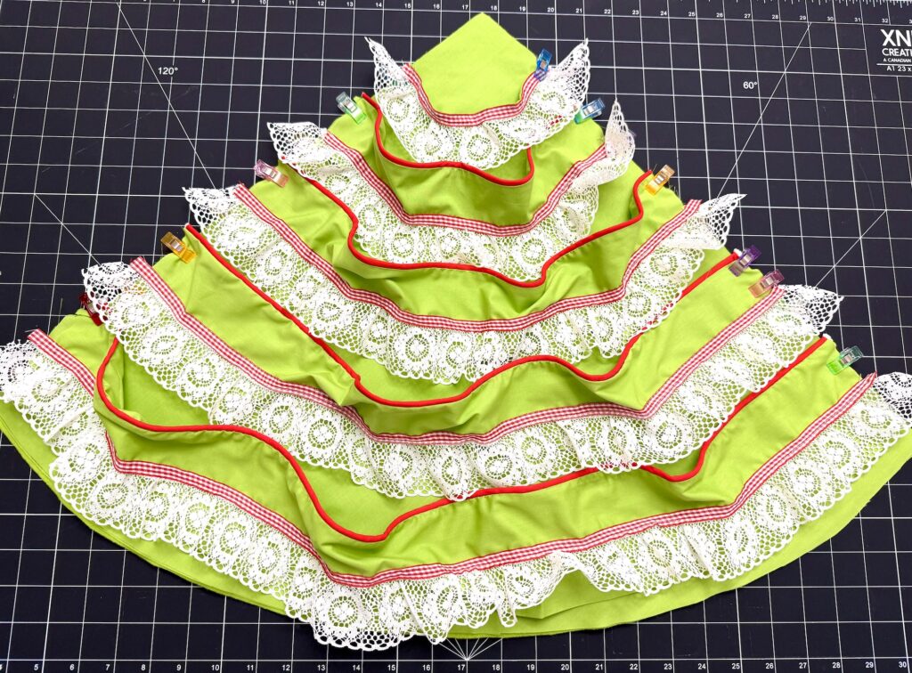 A green, tiered fabric advent tree with red trim and white lace edges is laid flat on a black grid cutting mat, secured with colorful clips along the top edge.