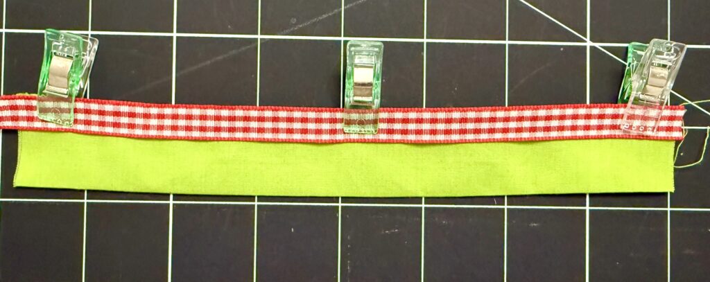 A strip of green fabric with a red and white gingham ribbon on top, held together by three sewing clips on a gridded cutting mat—perfect for crafting an advent tree.