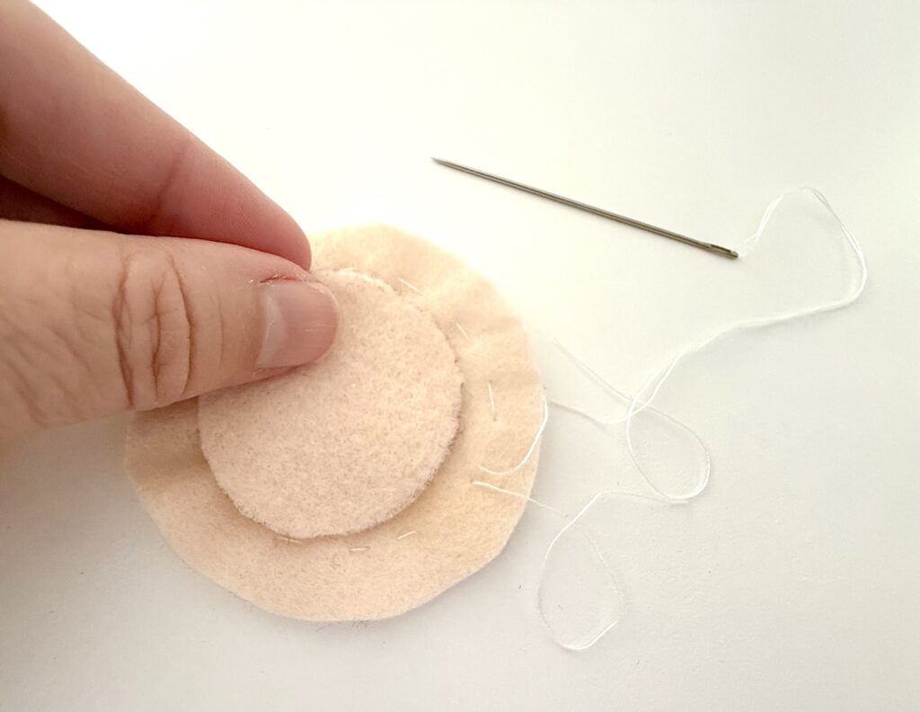 A hand holds two pieces of beige felt being sewn together with white thread; a sewing needle lies on a white surface nearby.