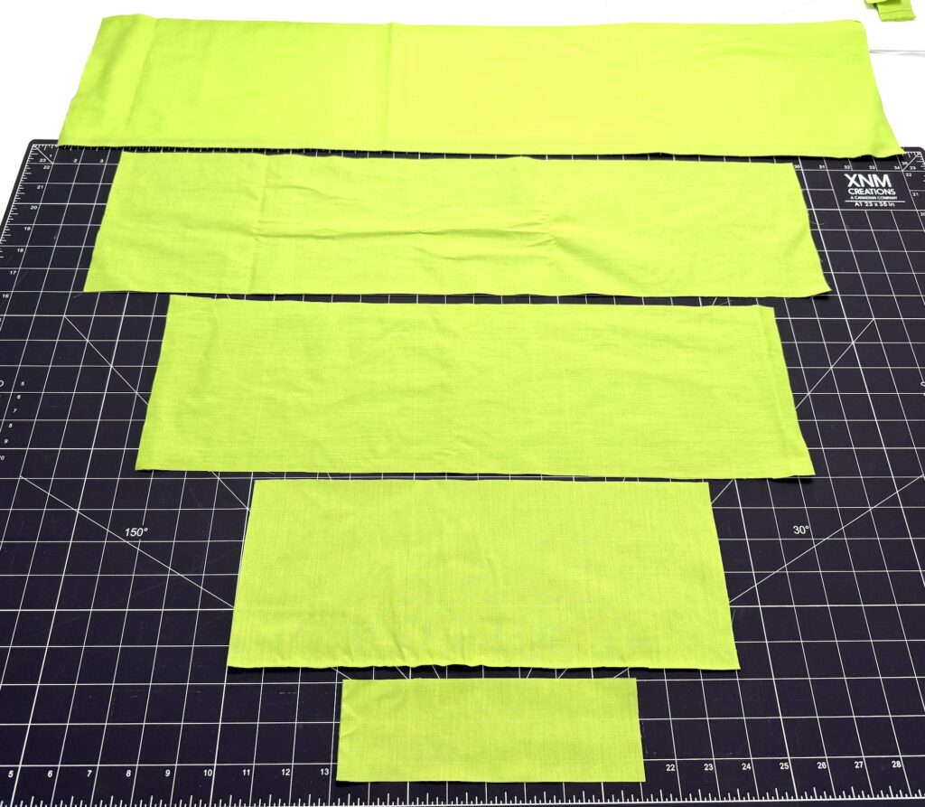 Four lime green fabric rectangles of varying sizes, perfect for crafting an advent tree, are laid out on a black gridded cutting mat with measurement markings.