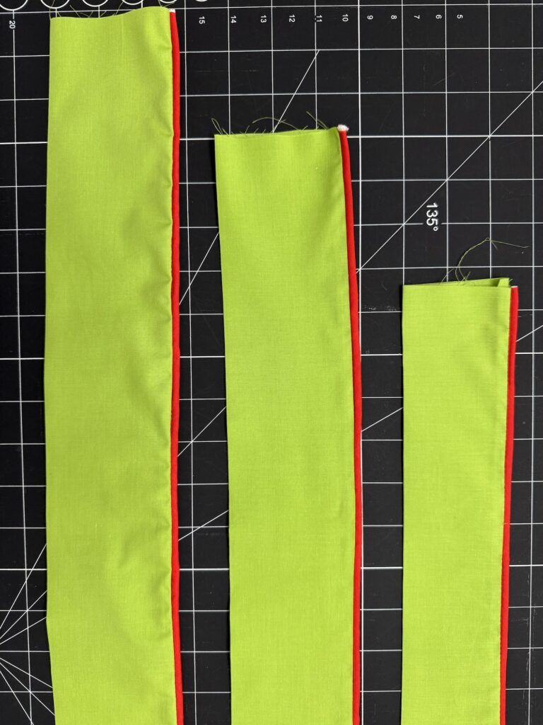Three green fabric strips with red piping edges, perfect for an advent tree project, are laid out vertically on a black grid cutting mat with white measurement markings.