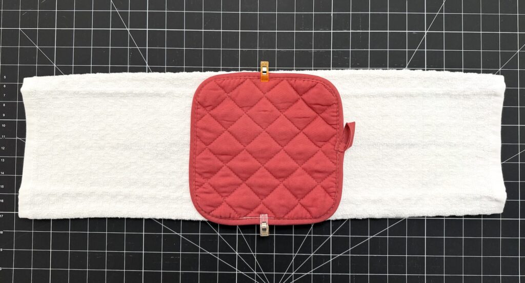A red quilted pot holder is clipped to the center of a white towel, both placed on a black grid cutting mat.
