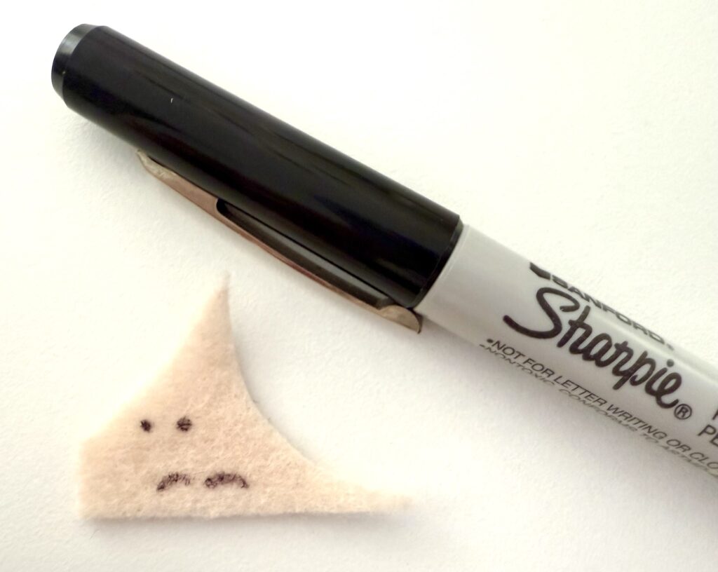 A black Sharpie marker next to a small triangular piece of beige felt with a sad face drawn on it.