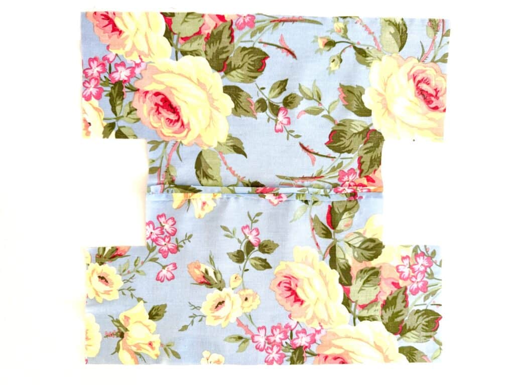 A piece of light blue fabric with a floral pattern, featuring large yellow and pink flowers, is cut into a specific shape with notches on two sides—perfect for making a tissue box cover.