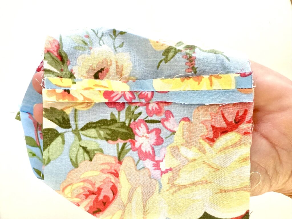 A hand holds a piece of light blue floral fabric with a seam folded and pressed open along the edge, ready to be sewn into a charming tissue box cover.