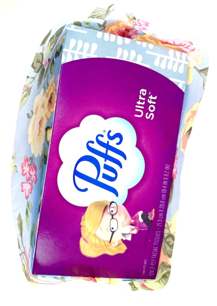 A box of Puffs Ultra Soft tissues sits in a floral tissue box cover, viewed from above on a white background.