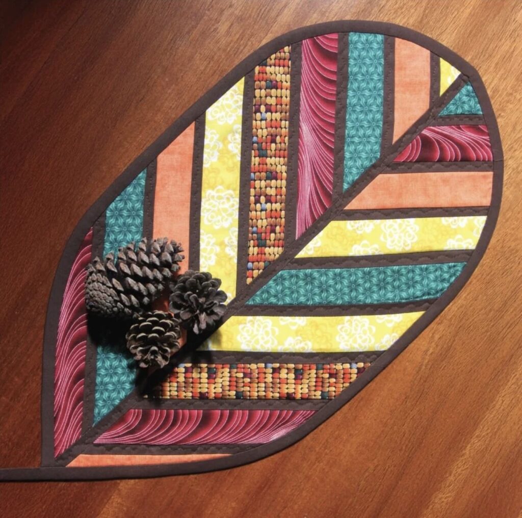 A quilted, leaf-shaped fabric placemat with colorful geometric patterns, displayed on a wooden surface with three pine cones on top.