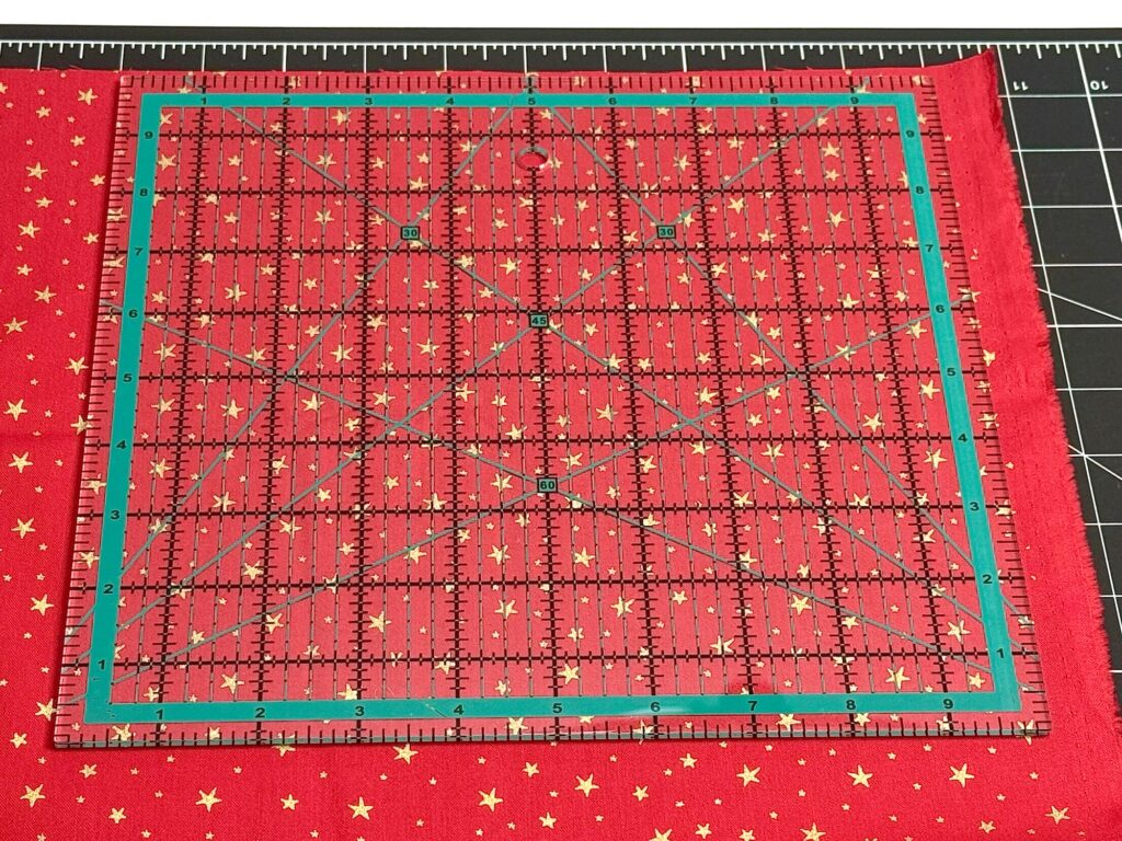 A clear quilting ruler placed on red fabric with gold stars, aligned on a black cutting mat marked with white grid lines and measurements.
