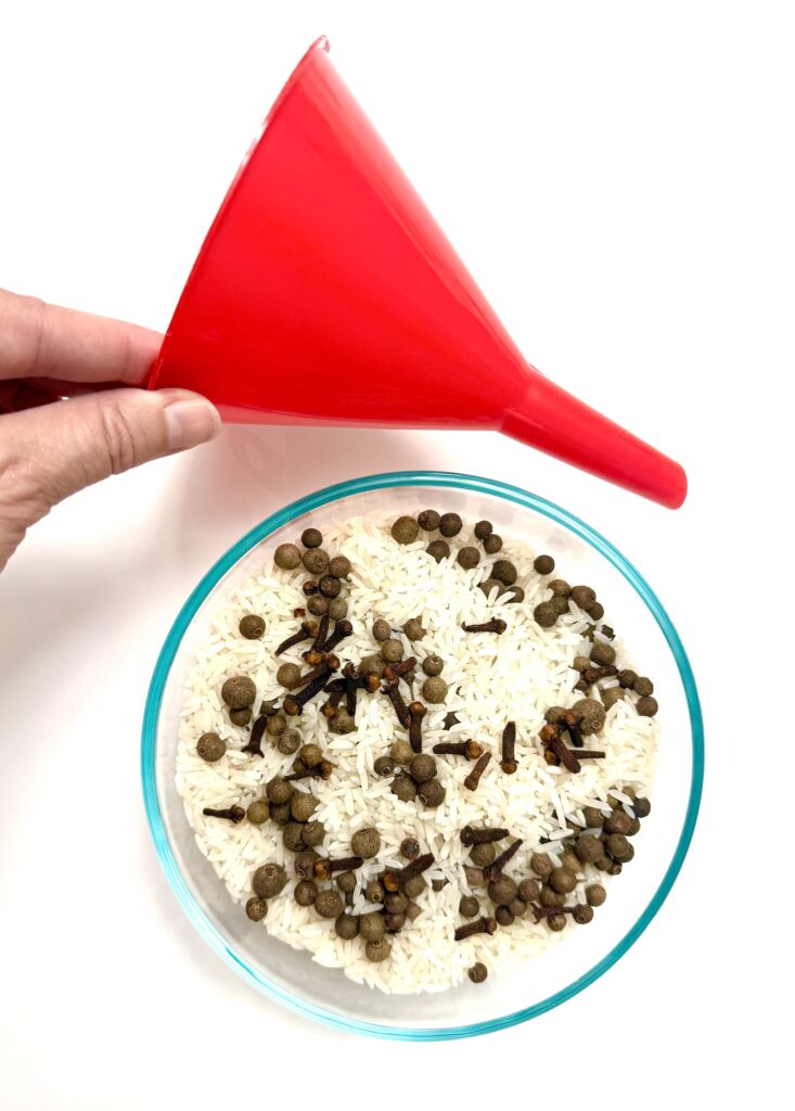 A hand holds a red plastic funnel next to a clear bowl containing white rice mixed with whole cloves and peppercorns on a white surface.