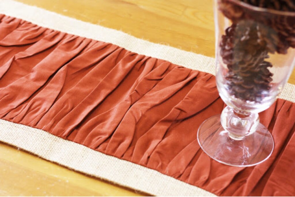 A close-up of an orange ruched fabric table runner on a burlap base, with a clear glass vase holding pinecones on a wooden table.