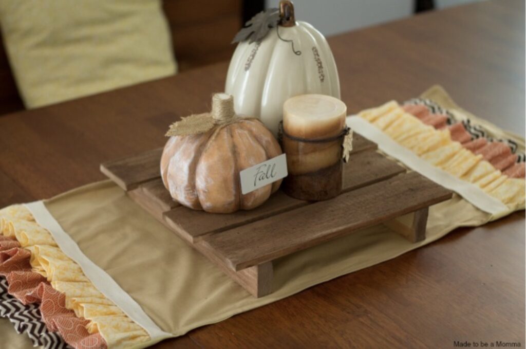 A wooden table centerpiece with two decorative pumpkins, a stacked candle, and a small “fall” sign on a wooden tray atop a ruffled fabric table runner.