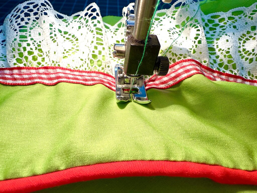 A sewing machine stitching green fabric for an advent tree with red trim and white lace, showing a close-up of the needle and presser foot in action.
