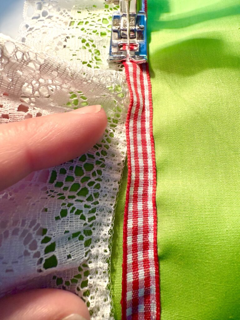 A close-up of a sewing machine attaching white lace and red gingham ribbon to green fabric, with a hand guiding the materials—perfect details for crafting an advent tree.